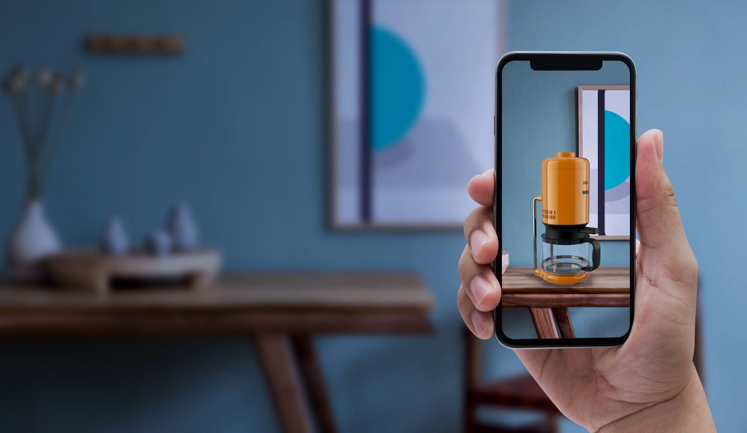 A hand holds a smartphone framing the empty top of a table. Thanks to Augmented reality the smartphone displays the table with on top a coffe maker digitally added.